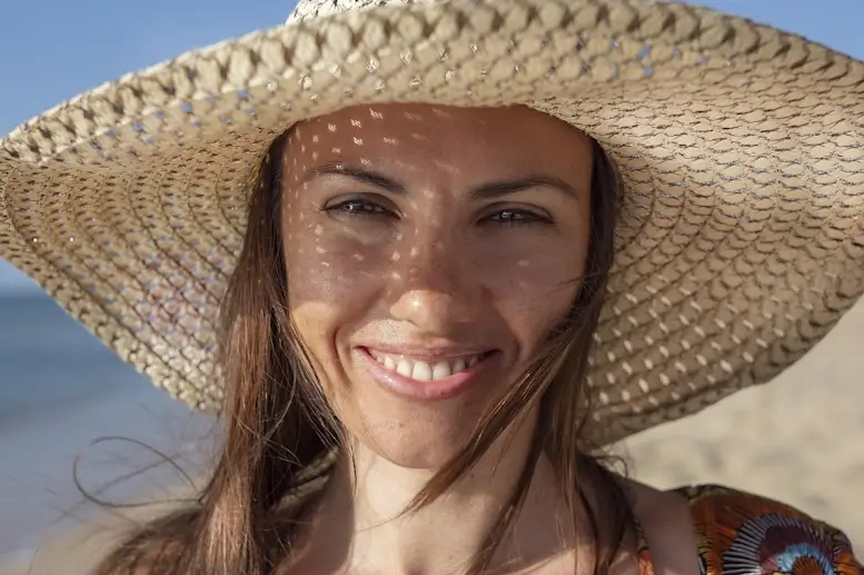 Une femme au large chapeau sourit sur une plage.